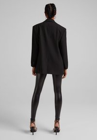 Svart skräddarsydd kavaj med en avslappnad passform, i kombination med blanka svarta leggings och sandaler med hög klack och remmar, sedd bakifrån.