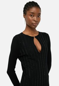 Abito lungo nero a maniche lunghe realizzato in tessuto a costine, con scollatura a V profonda e chiusura con bottone, impreziosito da sottili righe decorative.