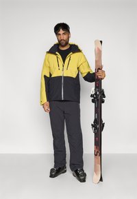 Conjunto de ski com uma jaqueta amarela e azul marinho com fechos de correr e bolsos, calças pretas e botas de ski pretas, segurando um par de esquis.