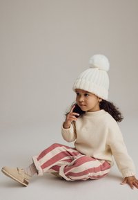Chapeau en tricot crème avec pompon, sweat-beige et pantalon à rayures roses. Chaussures beiges avec une sangle. Cheveux bouclés. Fond neutre.