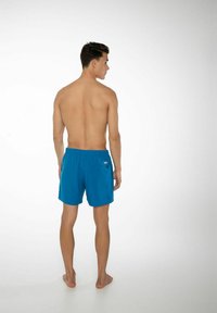 Blå simshorts med elastiskt midjeband, ett enda bakfack och en slät, lätt textur, sedd bakifrån.
