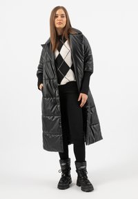 Long manteau d'hiver noir avec une finition brillante, doté d'un col châle, de boutons-pression et de poches latérales, porté par-dessus un pull en argyle noir et blanc.