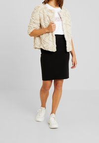 Beige keinoturkkinen takki, jossa on teksturoitu aaltoileva kuvio, päällä valkoinen t-paita, jossa on punaiset kirjaimet, sekä musta kynähame. Valkoiset tennarit.