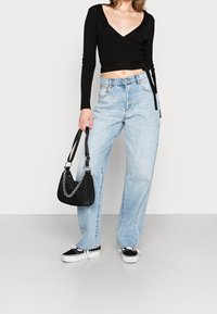 Frau trägt einen schwarzen Wickel-Pullover, hellblaue hochgeschnittene Jeans, schwarz-weiße Turnschuhe und hält eine schwarze Handtasche mit einer Kettenriemen.