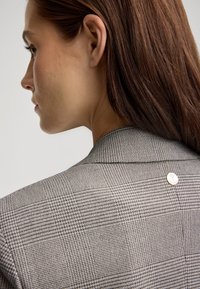 Gecontroleerde blazer met een subtiele grijs en beige patroon, voorzien van een notch-kraag en een metallic knoopdetail op de achterkant. Soepele textuur.