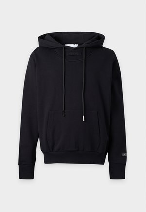 Homme portant un sweat à capuche noir, un jean bleu clair à jambes larges, des baskets rouges et des lunettes de soleil noires, debout sur un fond uni.