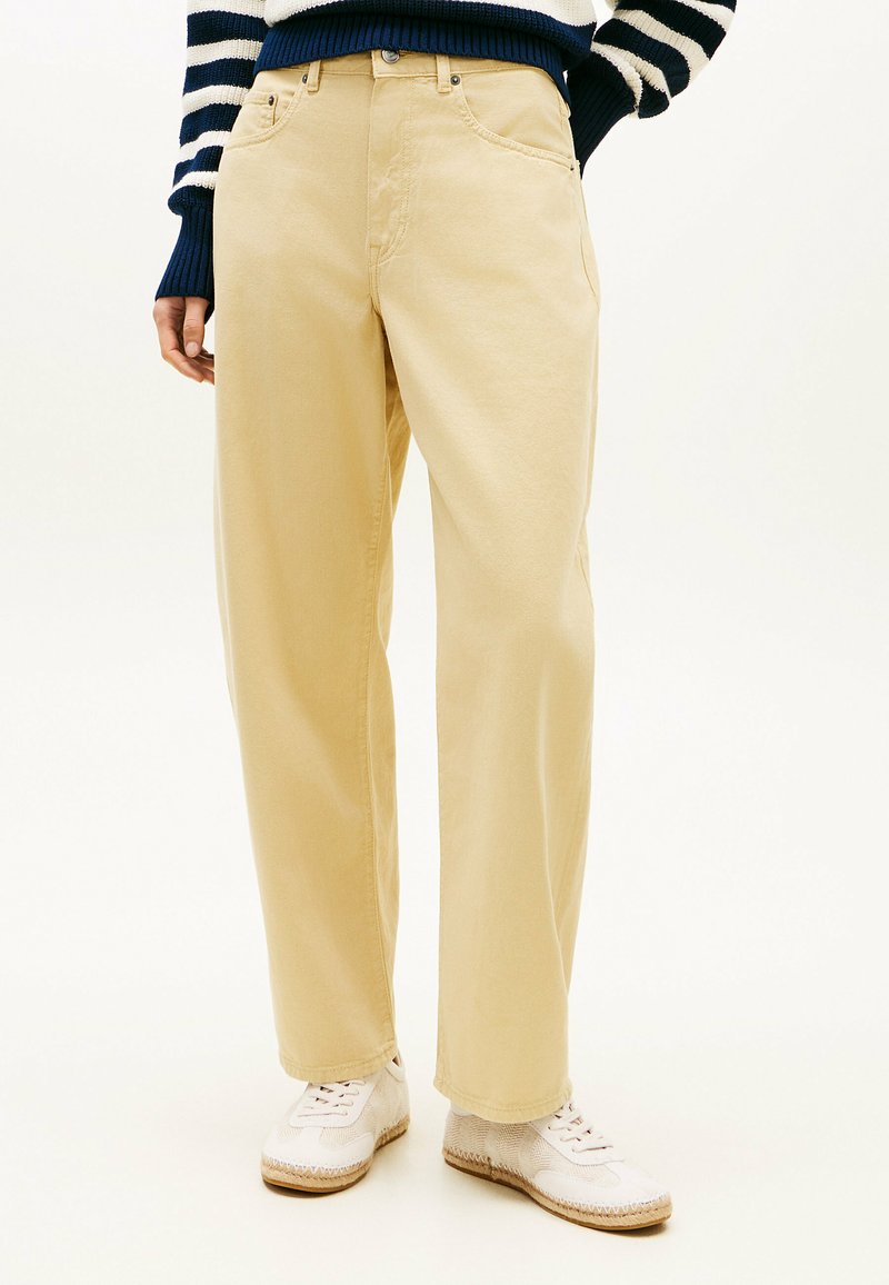 Personne portant un pantalon beige à jambes larges, des baskets blanches et un pull à rayures navy et blanc, les mains dans les poches, sur un fond uni.