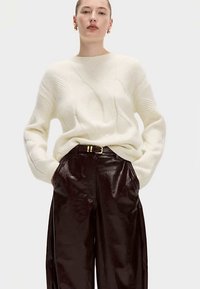 Krémový svetr s copánkovým vzorem a kulatým výstřihem, kombinovaný s lesklými tmavohnědými koženými kalhotami s vysokým pasem. Jednoduché zlaté kovové doplňky.