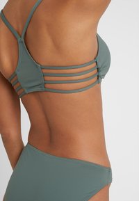 Grünes Bikini-Set aus strukturiertem Stoff, mit drei horizontalen Trägern am Oberteil und einem schlichten, tiefgeschnittenen Unterteil in passender Farbe.