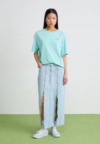 Tricou oversized de culoare verde mentă cu un logo grafic, purtat împreună cu o fustă maxi din denim deschis, care are două fermoare și slituri laterale. Adidași albi.