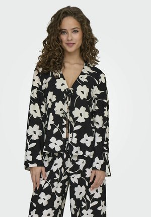 Schwarze Bluse mit floralen Mustern in Weiß, langen Ärmeln, einem V-Ausschnitt und einem Bindeband vorne. Kombiniert mit passenden Hosen.