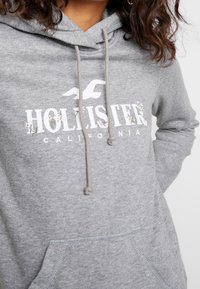 Närbild av person som bär en ljusgrå Hollister-hoodie med blommönster på logotypen och dragsnören som hänger ner.