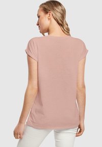 Camiseta de manga corta ajustada en suave tela rosa, con cuello redondo y puños enrollados, con una parte posterior lisa y un dobladillo ligeramente más largo.