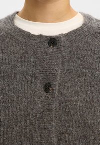 Šedý vlněný cardigan s texturovaným pletením, zdobený dvěma černými knoflíky a kulatým výstřihem. Látka působí měkce a mírně načechraně.