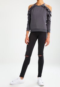 Sudadera gris oscuro con diseño de hombros descubiertos, acentos de volantes y mangas largas. Combinada con jeans skinny negros desgastados y zapatillas deportivas blancas.