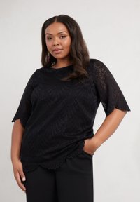 Top in pizzo nero con orlo a scallop e maniche corte, caratterizzato da un motivo a foglia. Il modello lo indossa con pantaloni neri, mani nelle tasche.