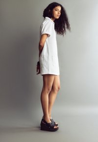 Witte denim korte jurk met korte mouwen en twee voorzakken, gecombineerd met zwarte platform sandalen met metallic accenten.