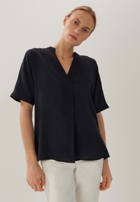 Schwarzes kurzärmliges Hemd mit V-Ausschnitt und Faltendetail, in lässiger Passform und glatter Textur, kombiniert mit hellen Hosen.