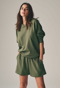 Zelený bavlnený oversized sveter s dlhými rukávmi, doplnený o voľné šortky v rovnakom odtieni. Obe kusy majú jemnú štruktúru a uvoľnený strih.