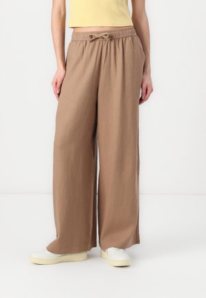 Kvinde iført beige vidde bukser med snøre i taljen og hvide slip-on sko, der står mod en ensfarvet hvid baggrund.