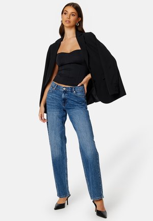 Haut noir sans bretelles, jeans en denim et talons hauts noirs. Le modèle porte un blazer noir surdimensionné drapé sur une épaule.
