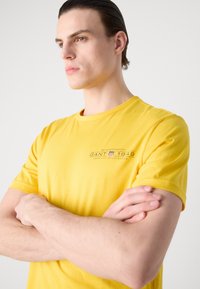 Kollane puuvillane T-särk ümmarguse kaeluse ja lühikeste varrukatega, millel on graafiline trükk vasakul rinnal, sile tekstuur.