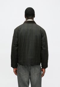 Ein Mann von hinten gesehen, der eine dunkle Jacke mit einem braunen Kragen und verblichenen grauen Jeans trägt, vor einem einfarbigen hellen Hintergrund.