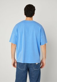 Modra bombažna majica s kratkimi rokavi in ohlapnim krojem, ki ima okroglo ovratnik in gladko teksturo. Obuta z jeans hlačami.