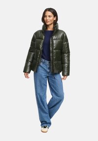 Vadderad, glansig grön puffjacka med hög krage, frontdragkedja och elastiska ärmmuddar, tillsammans med blå vida jeans och svarta sneakers.