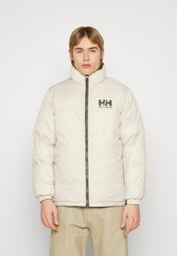 Švarkas pūkuotas šviesiai kreminės spalvos, su aukštu apykakle, užtrauktuku priekyje ir elastingomis rankovėmis. Ant krūtinės yra tamsiai žalia Helly Hansen logotipas.