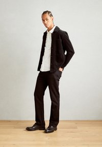 Veste en velours côtelé marron sur une chemise blanche, associée à un pantalon noir et des chaussures marron. Design à jambe droite, tissu texturé avec deux poches avant.