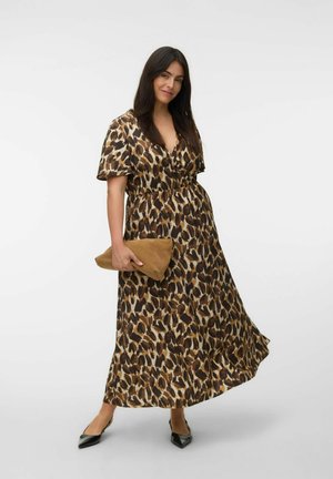Femme portant une robe longue imprimé léopard marron et noir, tenant une pochette beige, avec des ballerines noires pointues, debout devant un fond blanc uni.