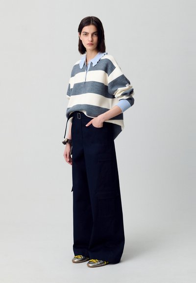 Maglione di grandi dimensioni lavorato a maglia con strisce grigie e bianche, camicia con colletto blu sotto. Abbinato a pantaloni larghi blu navy e sneakers metalliche.