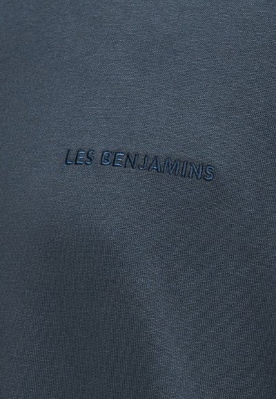 Σκούρο μπλε ύφασμα με κεντημένο το κείμενο "LES BENJAMINS" με νήμα σε αντίστοιχο μπλε χρώμα στο κέντρο της επιφάνειας.