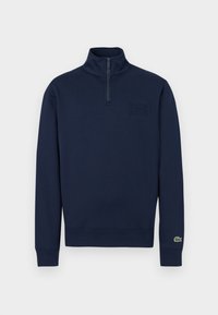 Sweat-shirt marine à demi-fermeture éclair avec col montant, poignets côtelés, petit logo embossé sur la poitrine et logo crocodile vert sur le poignet de la manche gauche.