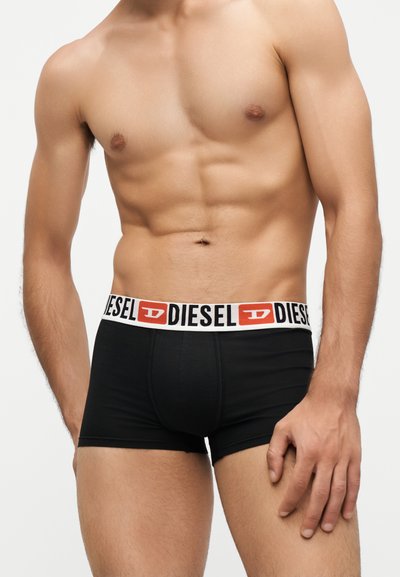 Мускулест мъж, носещ черни боксерки Diesel с бял колан, на който е логото на Diesel в черно и червено.