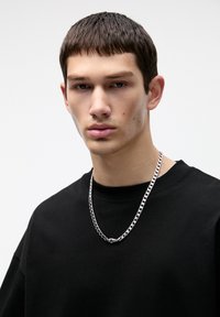 Tricou din bumbac negru cu guler rotund, asociat cu un colier din argint cu link-uri ovale și un detaliu central de închidere.