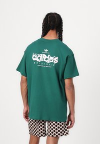Zaļš kokvilnas T-krekls ar lielu balto un rozā "adidas Originals" logo uz muguras. Brīvs piegriezums, īsas piedurknes, vienkrāsaina dizaina.