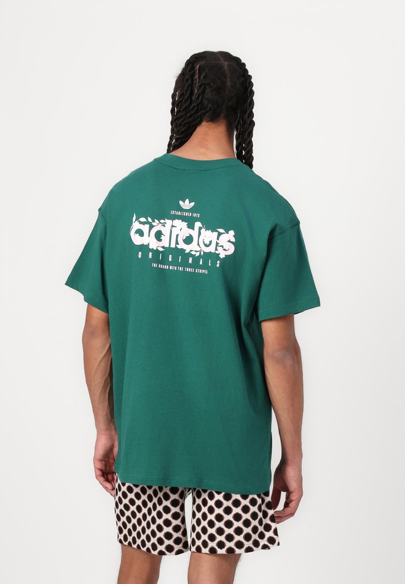 Zaļš kokvilnas T-krekls ar lielu balto un rozā "adidas Originals" logo uz muguras. Brīvs piegriezums, īsas piedurknes, vienkrāsaina dizaina.