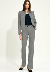 Kobieta stojąca w czarno-białym garniturze w pepitkę, z marynarką, dopasowanymi spodniami, czarną bluzką i czarnymi szpilkami na białym tle.