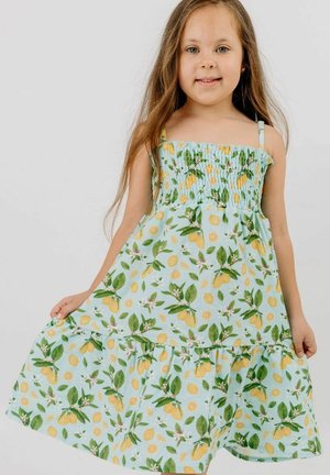 Junges Mädchen mit langen Haaren, das ein ärmelloses blaues Kleid mit einem Muster aus gelben Zitronen und grünen Blättern trägt und sanft den Rocksaum festhält.