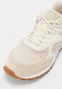 Beige sneaker dengan detail jala dan suede, tali putih, label lidah bermerek, dan sol karet gum di latar belakang putih.
