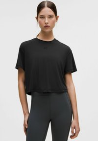 Fekete, rövid ujjú, rövid fazonú póló kerek nyakkivágással, finom logóval a mellkason. Magas derekú szürke leggingsszel párosítva.