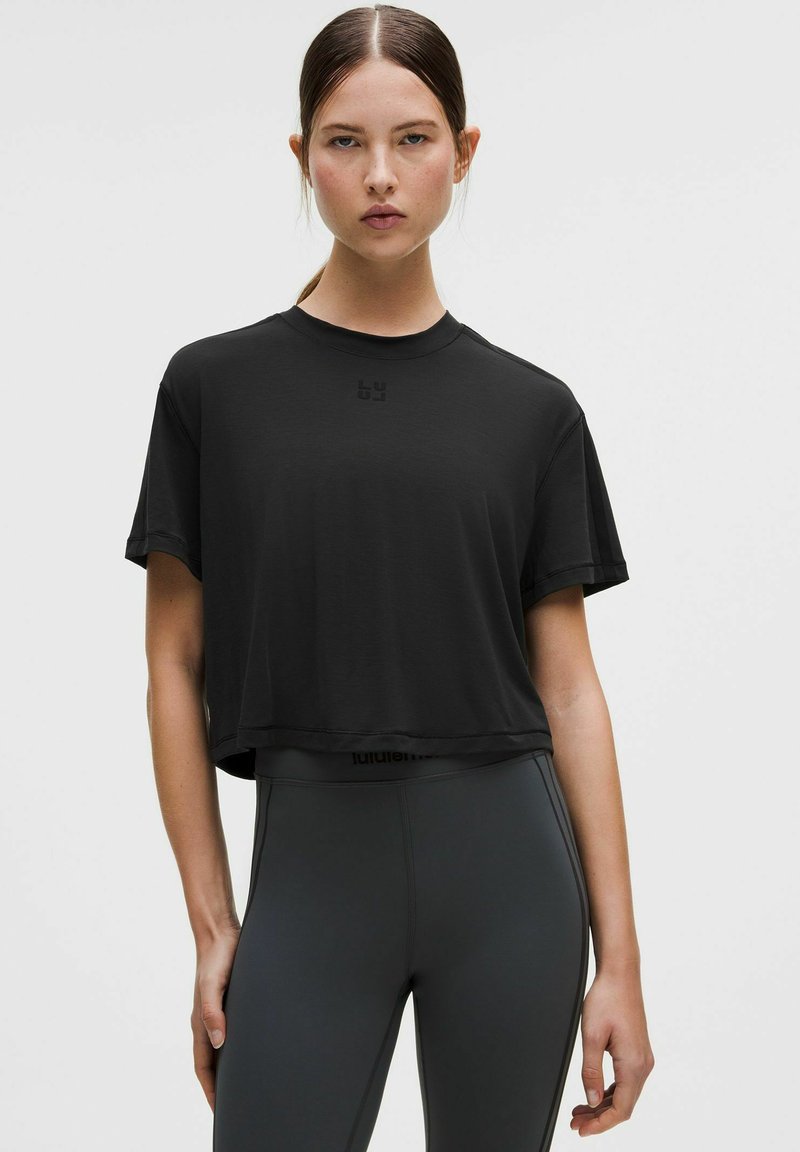 Fekete, rövid ujjú, rövid fazonú póló kerek nyakkivágással, finom logóval a mellkason. Magas derekú szürke leggingsszel párosítva.