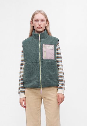 Jeune femme portant un gilet polaire vert avec une poche poitrine rose sur une chemise rayée à manches longues et un pantalon beige, debout sur fond blanc.