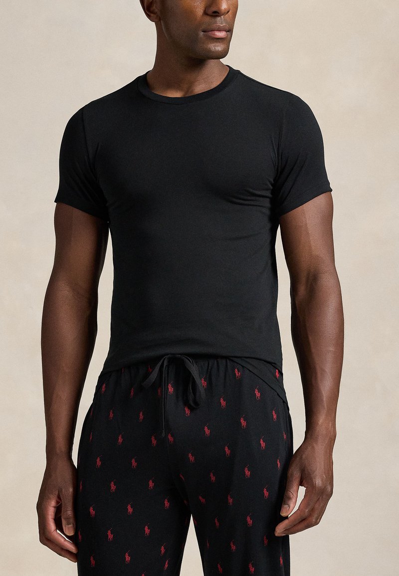 Homem vestindo uma camisa preta justa de manga curta e calças pretas com logótipos bordados de jogador de pólo em vermelho sobre um fundo bege.