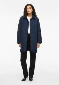 Modra prešita bunda z zapiranjem na gumbe, dolgimi rokavi in klasičnim ovratnikom; v kombinaciji z črnimi hlačami in špičastimi črnimi pete.