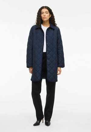Abrigo acolchado negro con cierre de botón, mangas largas y cuello clásico; combinado con pantalones negros y tacones negros de puntera.