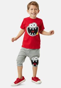 Punainen T-paita, jossa on monsteri-kasvojen graafinen kuva, harmaat shortsit, joissa on samanlainen design polvella, ja punaiset lenkkarit valkoisilla pohjilla.