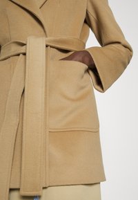 Beige wollen blendjas met een ceintuur in de taille, een opgestikte zak en wijde mouwen, voorzien van een zachte, gestructureerde afwerking en op maat gemaakte revers.